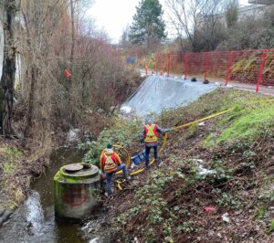 Utility locators scale a creek bank to inspect a raised manhole